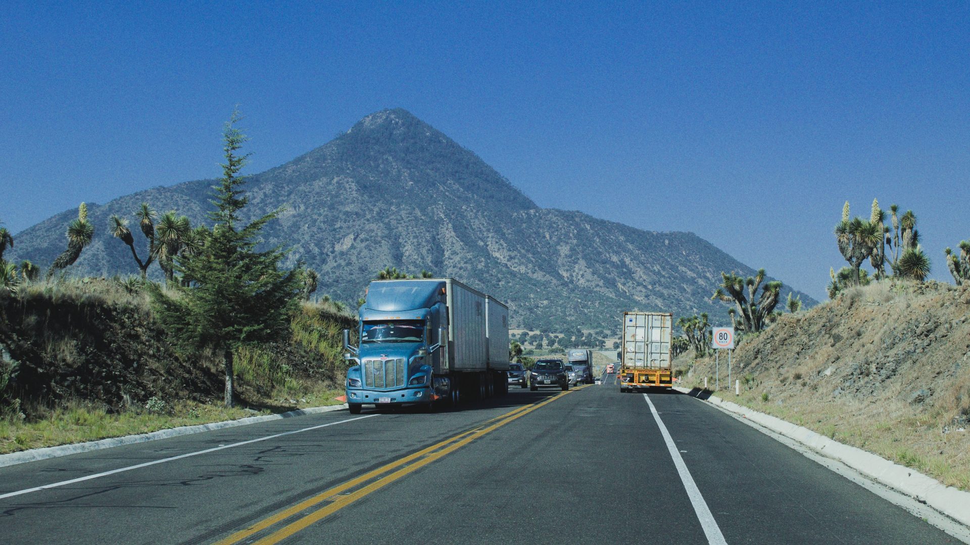 Trucks driving on a highway with a mountain backdrop under a clear blue sky.